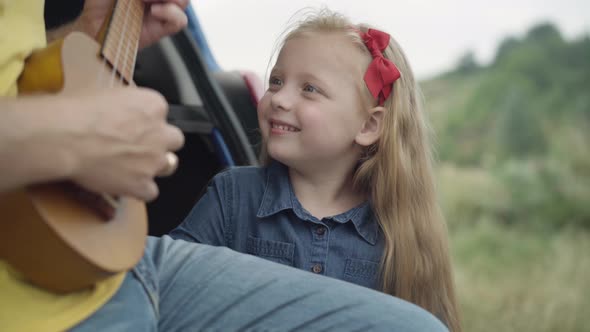 Portrait of Smiling Charming Cute Girl Admiring Unrecognizable Man Playing Ukulele Sitting in Car alt