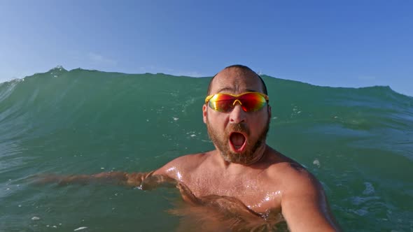 Active bearded man with yellow sunglasses having fun splashing with big sea waves breaking on his he alt
