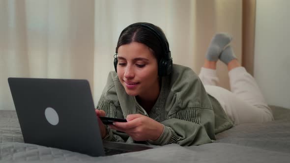 The Girl Lies on the Bed in Headphones Behind a Laptop with a Bank Card in Her Hands and Fills in