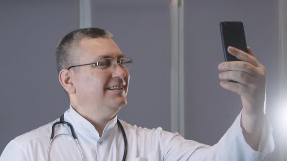 male doctor smiles and grimaces while taking a selfie on his phone alt