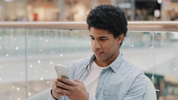 African Focused Millennial Man Sitting Indoors Swipes in Phone Screen Mobile Chat App Online alt