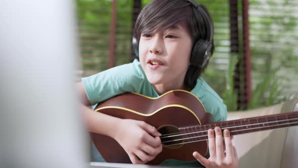 asian child boy online learning playing ukulele on armchair at living room