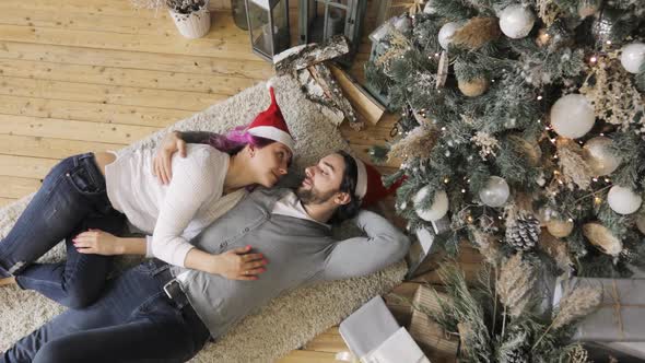 Young Couple in Love in Red Hats Lying Near Christmas Tree Hugging and Kissing.