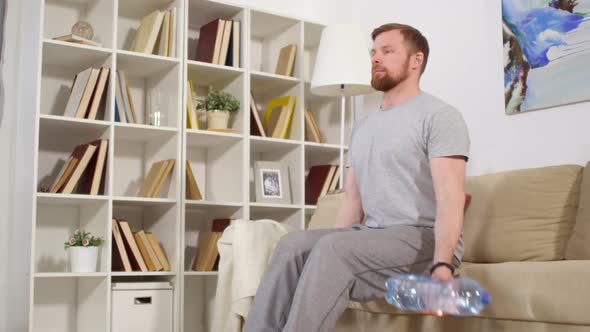 Man Sitting on Couch at Home and Doing Bicep Curls with Water Bottles alt