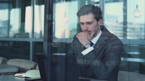 Young Businessman in a Suit Is Sitting on the Terrace of a Cafe and Working on a Laptop, View alt