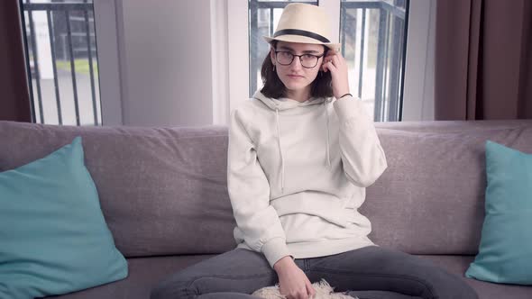 Girl in Glasses and a Hat Looks Straight Into the Camera Sitting Crosslegged on the Couch alt