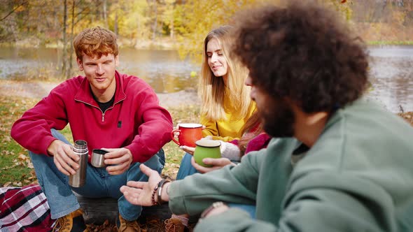Group of Young Men and Women Smiling and Enjoying Hot Tea From Thermos Sitting on Log in Autumn alt