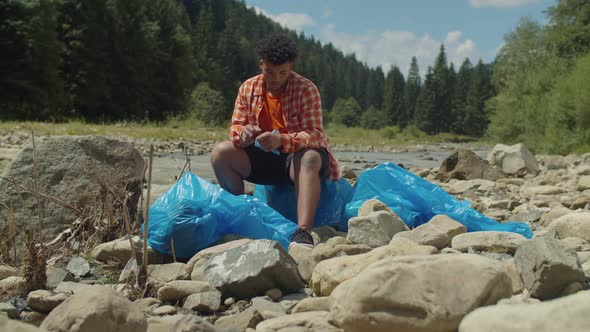 Portrait of Exhausted Black Man Ecologist Relaxing on Garbage Bag After Cleanup in Mountains alt