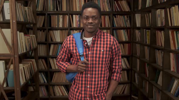 Black Male Student Holding Backpack in University or High School Library Smiling Looking at Camera alt