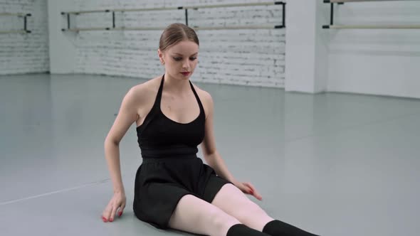 Elegant Ballerina Sitting and Stretching on the Floor of Dance Studio with Ballet Machines on the