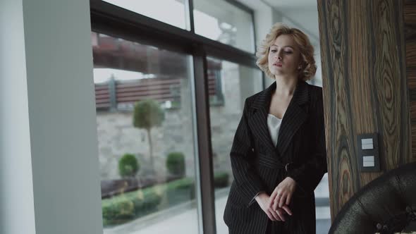 Pretty Model in Black Suit Leans on Wall and Crosses Hands alt