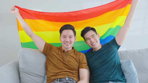 Portrait of Asian handsome man gay family hold LGBT flag and smile, look at camera in living room. alt