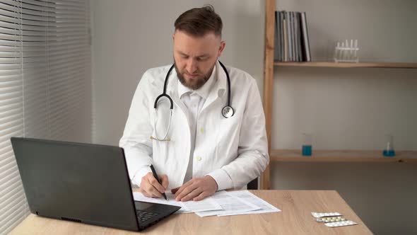 Serious Professional Male Doctor Using Laptop Computer in Hospital Office Watching Medical Webinar alt