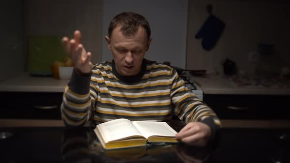 Young Man with Expression Reads a Book While Sitting at a Table in the Evening in the Kitchen alt