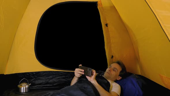 Relaxed Hiker in Sleeping Bag Using Smartphone Inside Open Tent, Alpha in