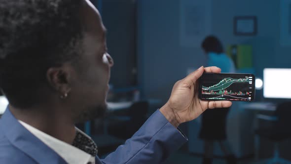 View Through Shoulder of AfricanAmerican Businessman Looking at Stock Exchange Graphs on Smartphone alt