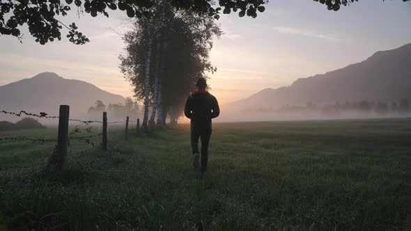 Man Walking Through Meadow Towards Foggy Landscape And Sunrise alt