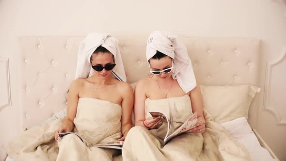 Two Beautiful Girls Lie on a Bed Under the White Blanket, Read Magazines and Surprised By Something