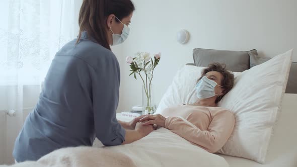 Home Caregiver With Face Mask Comforting Female Senior Patient Lying in Bed at Nursing Home
