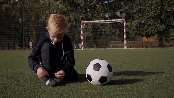 A Little Boy Soccer Player Ties His Shoelaces on a Football Field alt