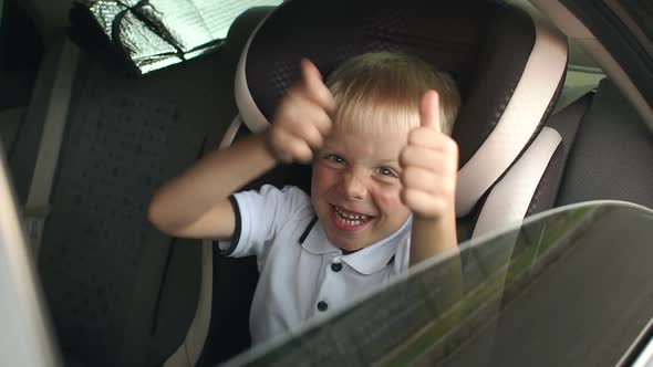 A Small Cheerful Boy Sits in a Brown Child Car Seat and Shows His Thumb Up alt