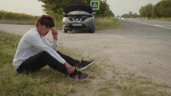 Worried Man Sits on the Grass Near His Broken Car alt