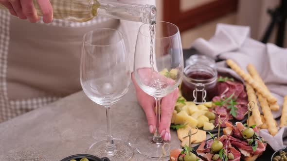 Woman Pouring Wine to a Glass From a Bottle at Domestic Kitchen with Meat and Cheese Plater on the alt