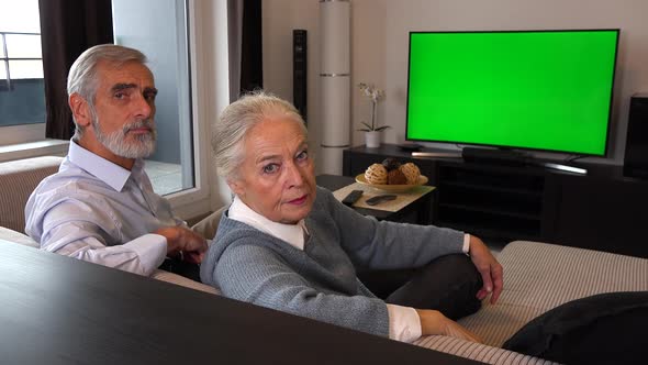 An Elderly Couple Watches Tv with A Green Screen and Shows Thumbs Down to The Camera alt