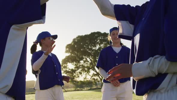 Diverse group of female baseball players on sunny pitch, taking a break to talk and high five alt