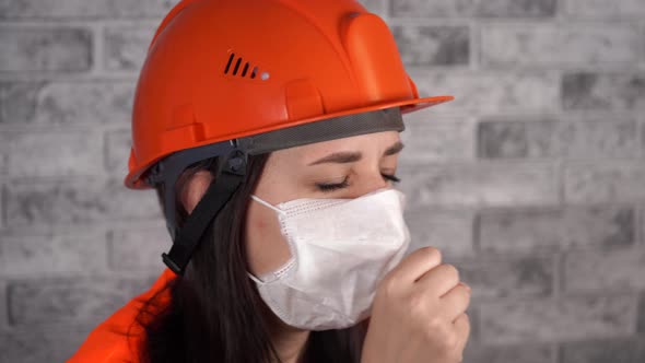 Female Construction Worker in Overalls and Medical Mask Coughing on Background of Gray Brick Wall alt