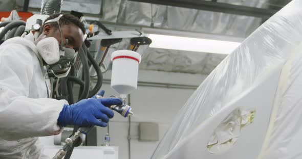 African American female car mechanic wearing protective clothes using a spraying gun to paint a car alt