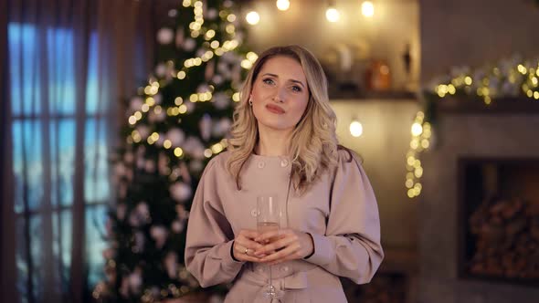Portrait of a Blonde in an Elegant Dress and with a Glass in Hand on the Background of a Living Room alt