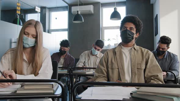 Young African American Student in Medical Mask Sits at Desk Among Multiethnic Classmates Raises Hand alt