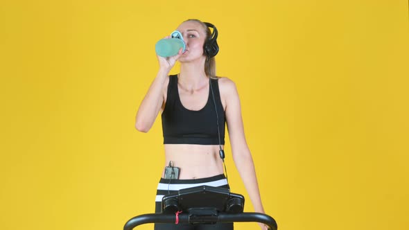 Fit blonde girl in sportswear with headphones works out on a treadmill alt