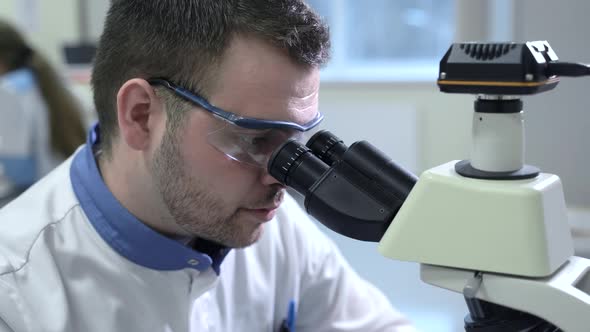 Portrait of Male Scientist Looking Through Microscope Sitting at Table in Pharmaceutical Lab Spbas alt