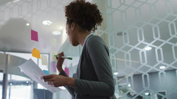 Mixed race businesswoman writing with green marker on memo note on transparent board in office alt
