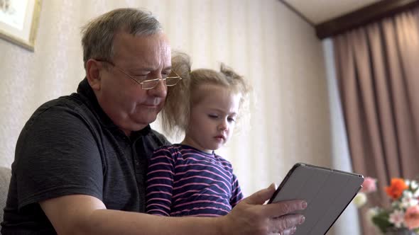 Granddaughter and Grandfather Are Sitting with a Tablet. The Girl Frowns at the Tablet. Sitting on alt
