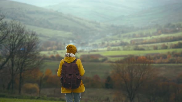 Woman Hiker with Backpack Walking By Green Meadow Among Hills Girl Wearing Yellow Jacket and Hat alt