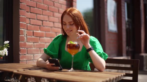 Portrait of Smiling Young Redhead Woman Wearing Green Dress Using Typing Cellphone Drinking Cocktail alt