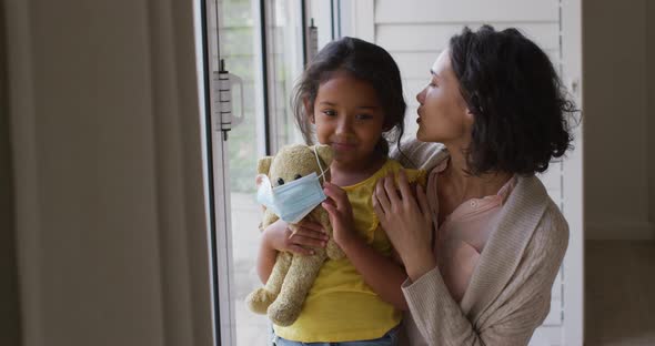 Hispanic mother and daughter embracing in window holding teddy bear with face mask alt