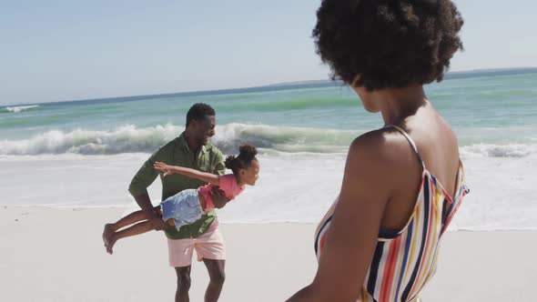 Smiling african american couple with daughter playing on sunny beach alt