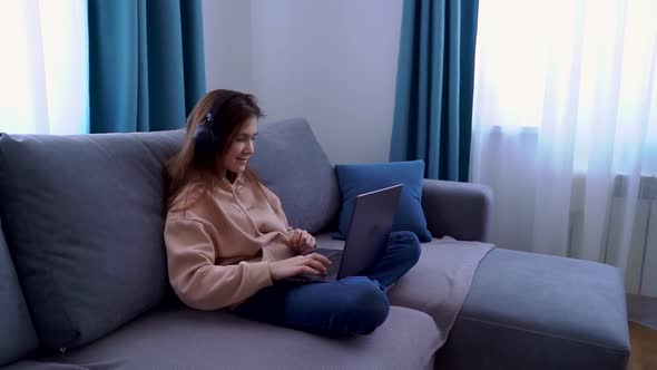 Smiling Young Girl in Headphones Using Laptop While Sitting on Sofa at Home alt