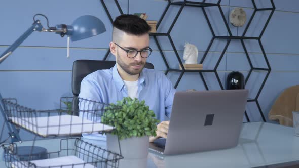 Young handsome man in glasses working on his laptop and sitting in the office at home alt