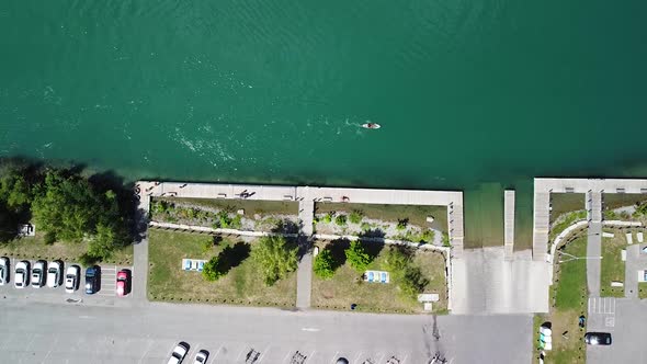 Aerial top down shot of parking cars beside river with person paddling on sup board during sunny day alt