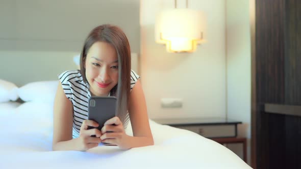Texting on Smartohone in Bed. A Young Happy Asian Woman in Bedroom Chatting With Mobile Phone, Slow alt