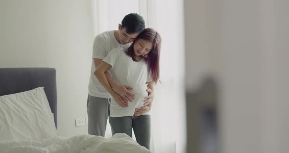 Happy Asian husband touching his wife's pregnant belly while standing beside a window.