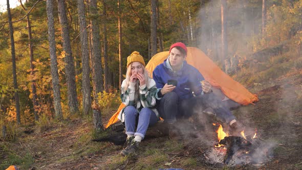 Young Couple of Tourists Camped in Woods Sit By Fire Eat and Meet the Sunset