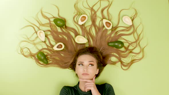 beautiful girl lying on green background with avocado fruits on long hair, woman thinking,choice  alt