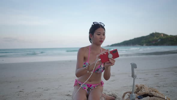 Beautiful Asian Girl Connecting a Phone to Osmo Mobile 5 OM5 Gimbal Sitting on Sandy Island Beach By alt