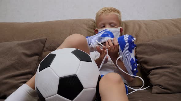 A Small Sad Boy in a Medical Mask Sits on a Sofa with a Soccer Ball and Cleats alt